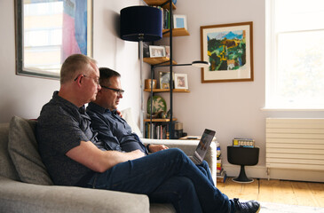 Older same sex male couple browsing laptop whilst sat on a sofa