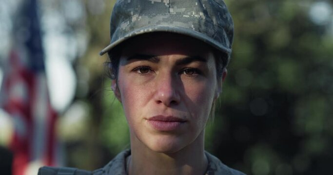 Close Up Slow Motion Portrait of Young Female Patriotic Soldier Standing in front of the American Flag and Looking at the Camera. Devoted Woman in Camouflage Military Uniform Ready to Serve and Defend