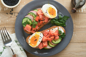 Healthy sandwich with bread or toast, smoked salmon, soft egg, cream cheese, cucumber, radish, black sesame and parsley on old wooden table. Delicious protein fish sandwich for breakfast. Smorrebrod.