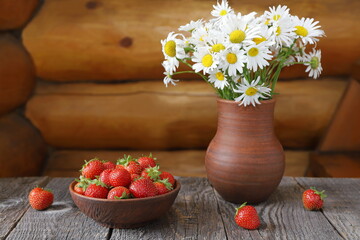 Obraz na płótnie Canvas A clay bowl of ripe strawberries next to a bouquet of field daisies in a pottery jug against a log wall.