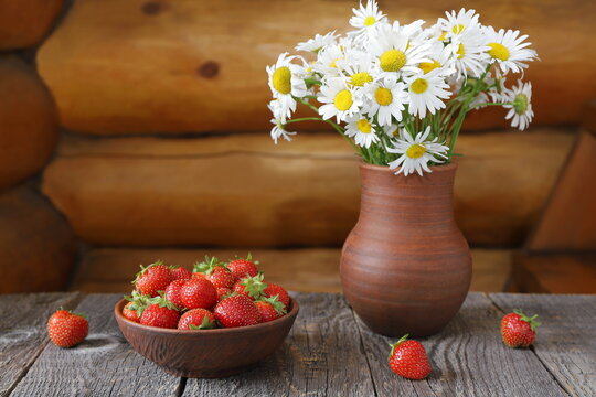 A clay bowl of ripe strawberries next to a bouquet of field daisies in a pottery jug against a log wall.