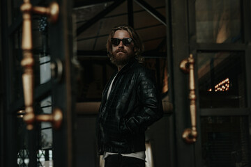 Fototapeta na wymiar Brutal caucasian man with long beard hipster in black leather jacket stay next to glass door