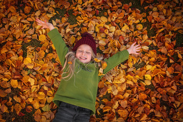 A little beautiful blonde girl in a burgundy hat and a green hoodie lies in red autumn leaves, top view