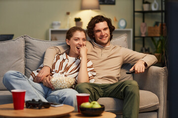 Portrait of happy young couple watching movies at home and eating popcorn sitting on couch together