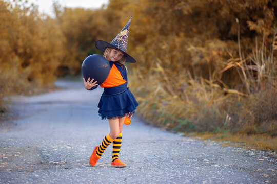 A girl in a witch costume for Halloween walks down the street with festive air hats in the form of pumpkins. Witch hat.