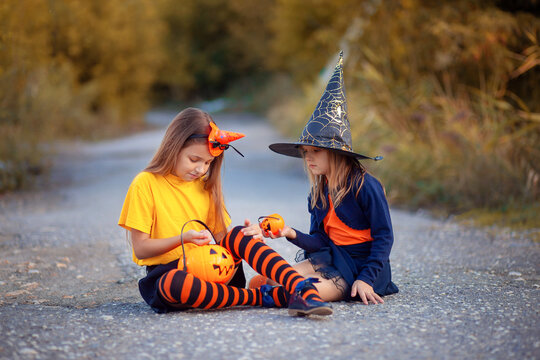 Two girls in Halloween costumes sitting on the asphalt on the street and sharing their candy. Children count candy collected for Halloween