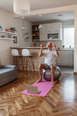 Man sitting on fitness ball, working out with dumbbells in his home gym, taking care of his health, exercising in living room. Active lifestyle concept.
