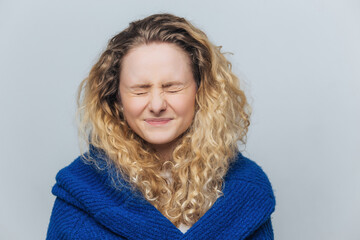 Young woman, wearing knitted sweater, anticipates gift with closed eyes, isolated on blue background.