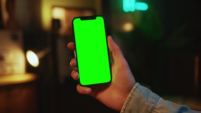 In the evening hands man holding and using mobile phone with a vertical green screen indoors. Social media. Mock up