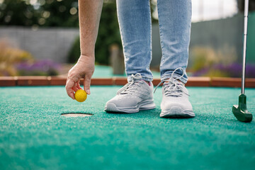 Woman plays mini golf and taking ball out from the hole after successful putting - obrazy, fototapety, plakaty