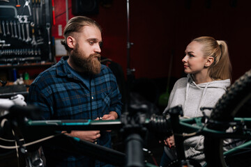 Obraz na płótnie Canvas Portrait of cycling repairman having conversation with blonde female client, talking about problem of bicycle detected during diagnostics in repair shop with dark interior. Concept of bike maintenance