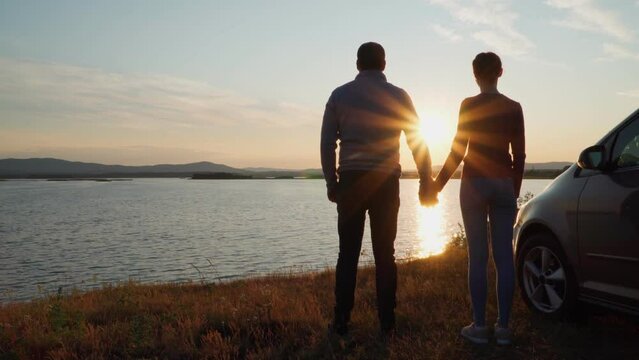 A man and a woman stand on the shore of the lake near their car and admire the sunset, holding hands. The golden hour.