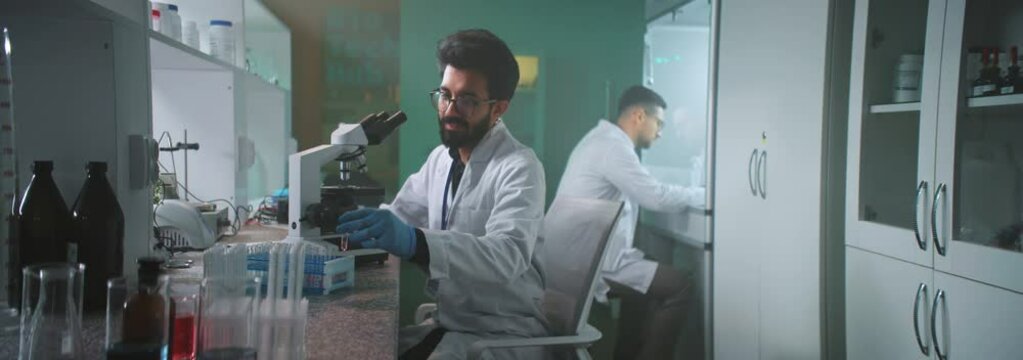A smiling man scientist with glasses and protective gloves looking under the microscope holds a tube in the laboratory. Medicine, biotechnology, chemistry. In the background colleague walking