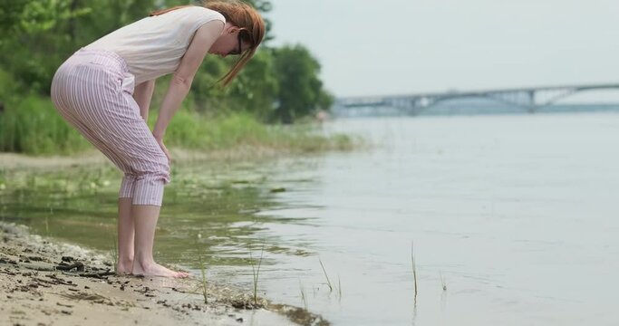 Young woman examines living creatures near river bank. Girl bent over water on shore, summer, river, bridge, green trees.