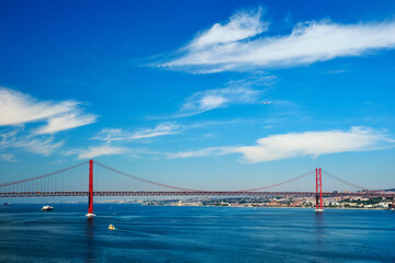 View of 25 de Abril Bridge famous tourist landmark of Lisbon connecting Lisboa to Almada on Setubal Peninsula over Tagus river with boats yachts, vessels and flying plane. Lisbon, Portugal