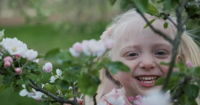 Video portrait of happy smiling round-faced girl with blond hair near flowering tree. Child is smiling all tooths.