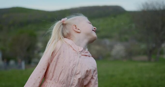 Fair-skinned blonde child girl in pink jacket on outside on green meadow while flower petals or snow are flying.
