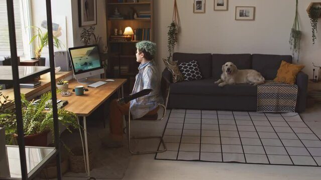Caucasian freelance girl with arm prosthesis sitting down at wooden desk against window and starting work on computer in bright cozy apartment