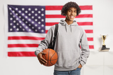 Young african american male with a backpack holding a basketball and smiling in front of USA flag