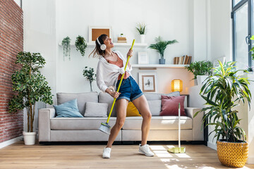 Funny motivated woman dancing and listening to music with headphones while sweeping the house