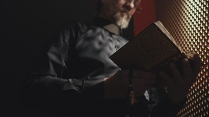 Low angle selective focus shot of mature Catholic priest holding rosary standing in confessional booth opening Bible book