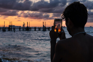 Back view silhouette of a young latin man taking a photo with a smartphone of a beautiful sunset at the caribbean beach