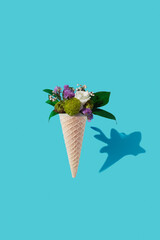 Waffle cornet with colorful flowers and green leaves on blue background. Flower ice cream, spring and summer concept with first flowers, mother's day, top view.