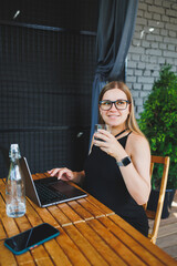 A young woman in glasses and a black dress sits on the summer terrace of a cafe and works at a laptop. Female freelancer works remotely online while sitting in a summer cafe.