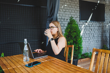 Fototapeta na wymiar A young woman in glasses and a black dress sits on the summer terrace of a cafe and works at a laptop. Female freelancer works remotely online while sitting in a summer cafe.