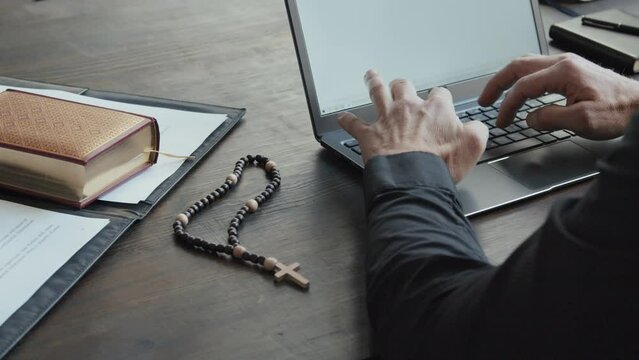 Unrecognizable Catholic priest sitting at wooden table with papers, prayer book and rosary on it typing something on laptop
