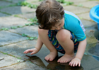 young boy in a bathing suit playing in a puddle of water in the backyard next to a shallow swimming pool
