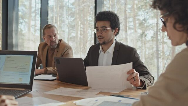 Medium shot with halo effect of young multiracial man in glasses sitting at table at team meeting on bright sunny day, with laptop and document, and making suggestion to colleagues