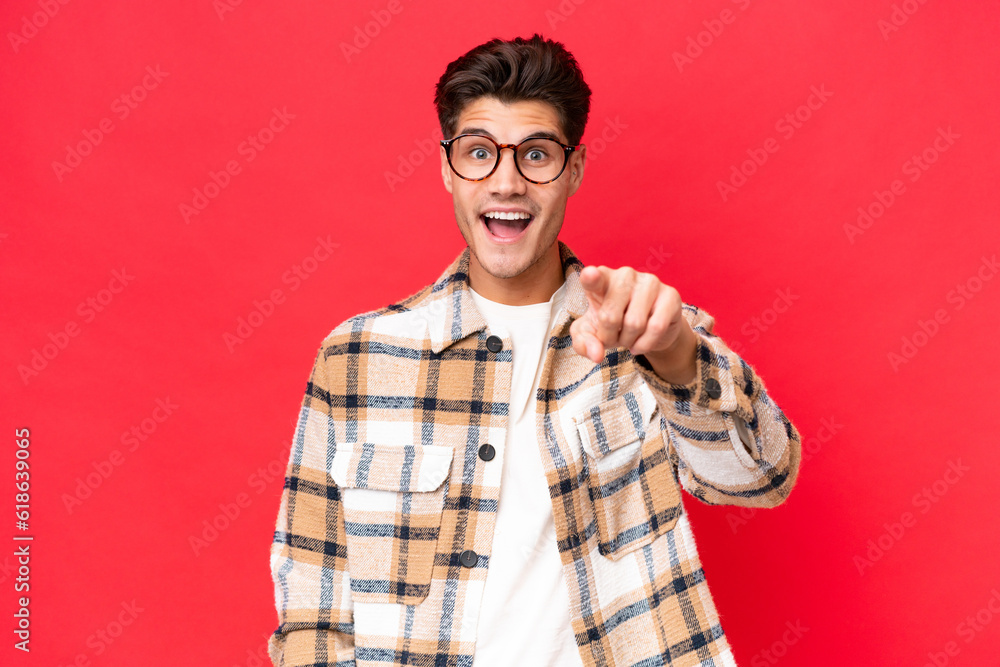 Wall mural young caucasian handsome man isolated on red background surprised and pointing front