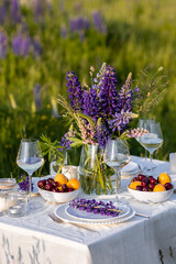 Romantic table decor for a loving couple on the blooming meadow with purple lupines. Two glasses of wine, flowers in a vase, silverware, fruits, wooden furniture and picnic basket. Sunset, golden hour