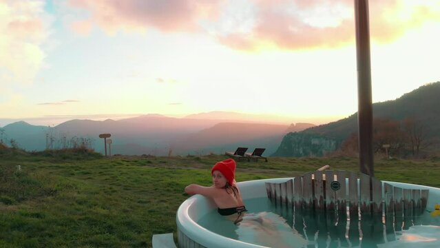 Drone shot unveils a picturesque scene. Young woman relaxes in natural-fed hot tub. Blue hot water and breathtaking spring sunset. Outdoor bath is epic concept for weekend getaway or holiday retreat.