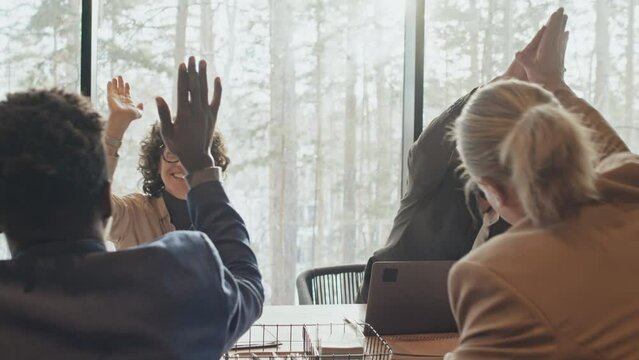 Medium shot with halo light of diverse startup team sitting at table at business meeting, man sharing great news, everybody jumping up, cheering and doing high fives