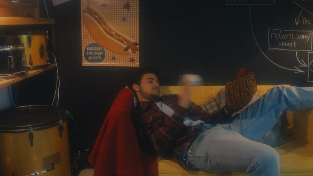 High angle view of young Middle Eastern man wearing baseball glove playing with ball while relaxing on couch, 90s aesthetics