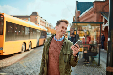 Mature man using a smart phone while waiting for his bus at the bus stop