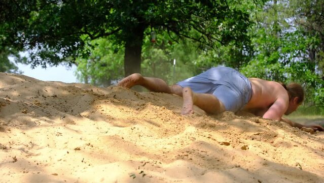Shirtless slightly obese young man crawling over pile of sand from left to right having fun after back yard pool party.