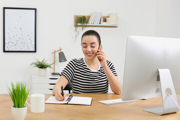 Home workplace. Happy woman with pen and notebook talking on smartphone at wooden desk in room