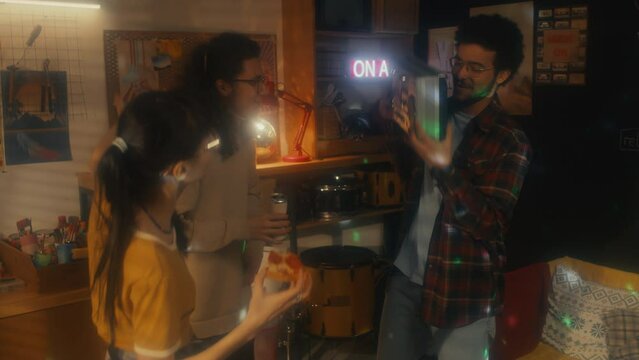 Group of three best friends eating pizza, drinking soda and dancing at party in garage, 90s aesthetics