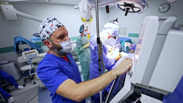 Experienced anesthesiologist pressing the buttons of medical equipment working in the surgery room. Surgeons at backdrop performing surgery.