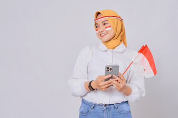 Excited young Asian Muslim woman wearing hijab holding mobile phone and Indonesian flag while celebrating Indonesian independence day on 17 August isolated over white background