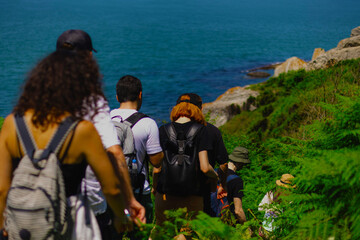 Group of hikers with backpacks wade through thickets of ferns along seashore on sunny summer day