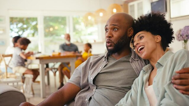 Shocked couple at home on sofa streaming horror show or movie to TV with multi-generation family in background - shot in slow motion