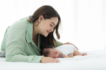 Happy Asian young mother takes care and kissing her newborn baby on white bed. Asian newborn baby sleeping in prone position with mother