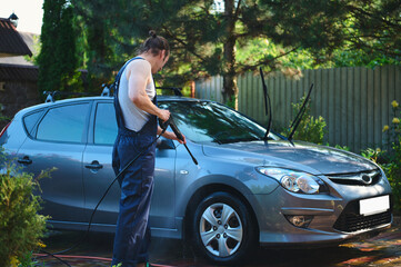 Full-length portrait. Car serviceman in blue work uniform, cleaning a vehicle with a high pressure water jet outdoors. High pressure water cleaner for manual car washing. People Technologies Transport