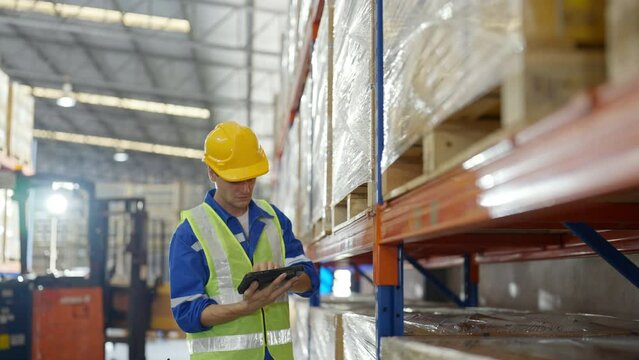 Young caucasian man warehouse workers holding digital tablet checking inventory management packaging boxes. Man staff wearing vest and safety helmet and walking count the box at storehouse