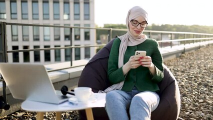 Full length view of charming woman in headscarf holding phone while sitting in beanbag near coffee table with gadgets. Delighted muslim person adoring city view in sunlight on panoramic terrace.