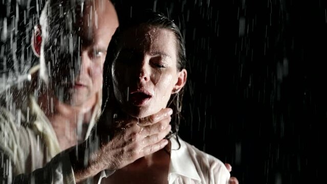 A brutal bald man in the pouring rain on a black background holds his beloved woman by the neck in loving caresses